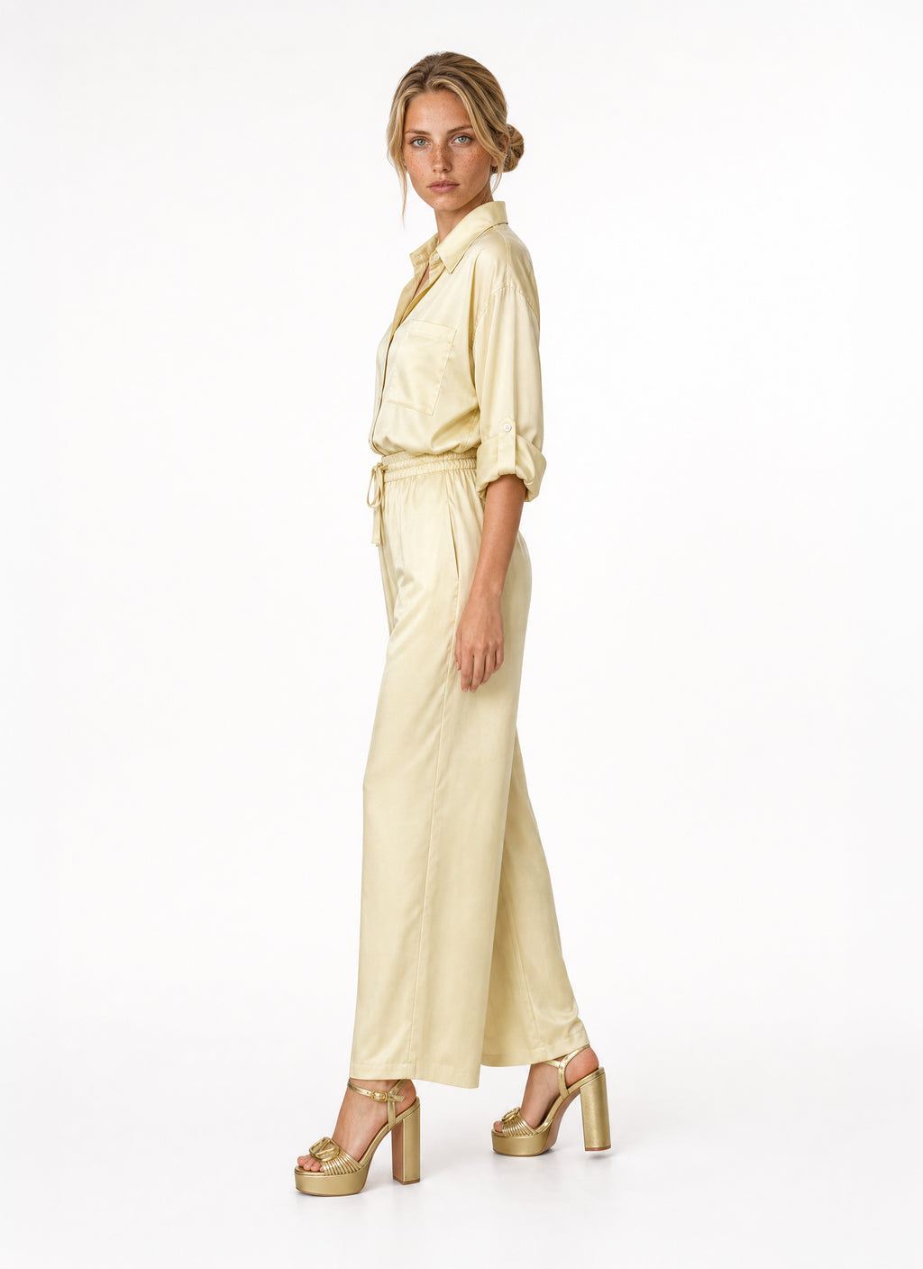 A woman modeling a pale yellow satin blouse and pant with a tie waist and wide legs, paired with metallic gold platform sandals, against a plain white studio background.