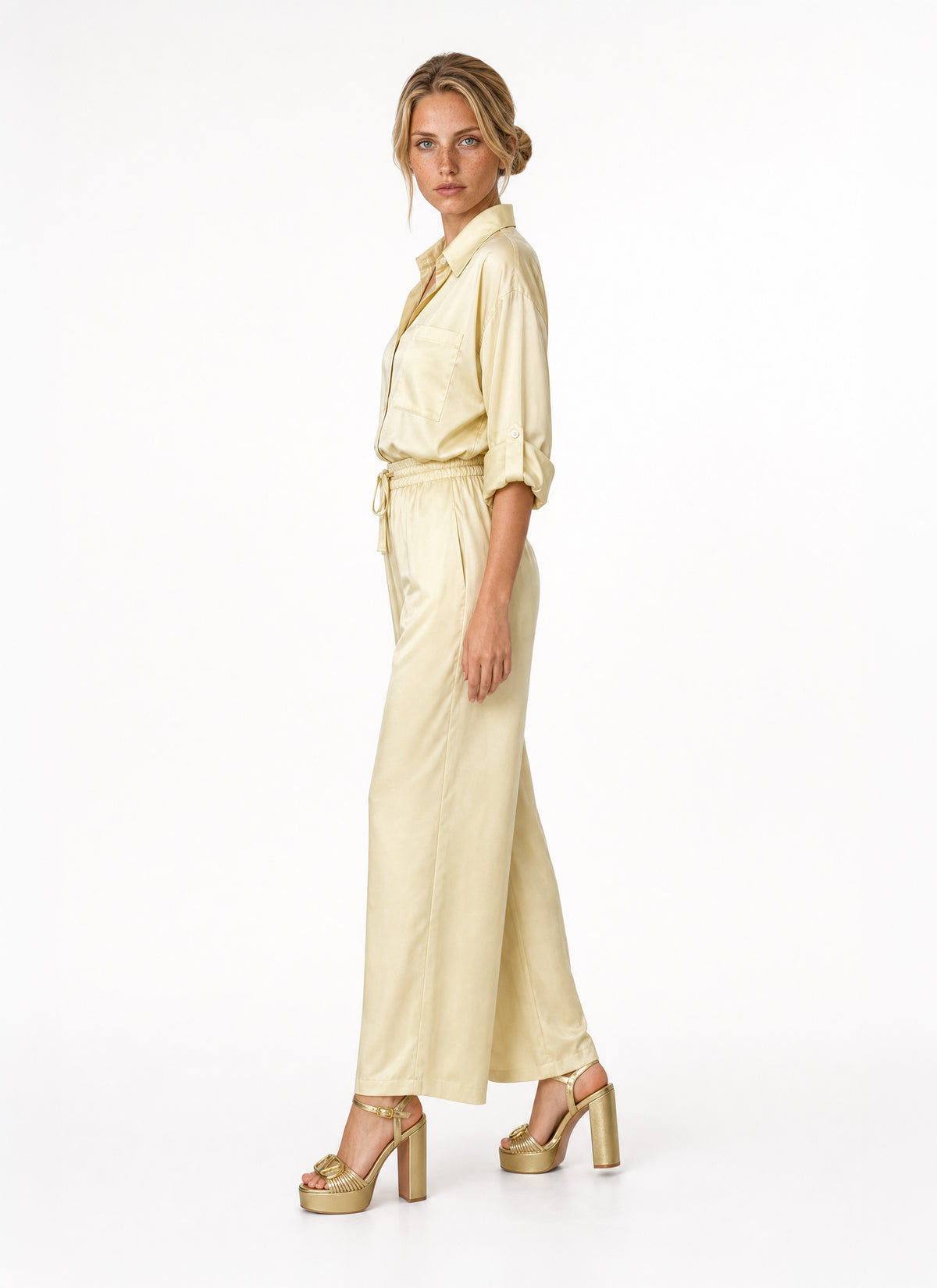 A woman modeling a pale yellow satin blouse and pant with a tie waist and wide legs, paired with metallic gold platform sandals, against a plain white studio background.