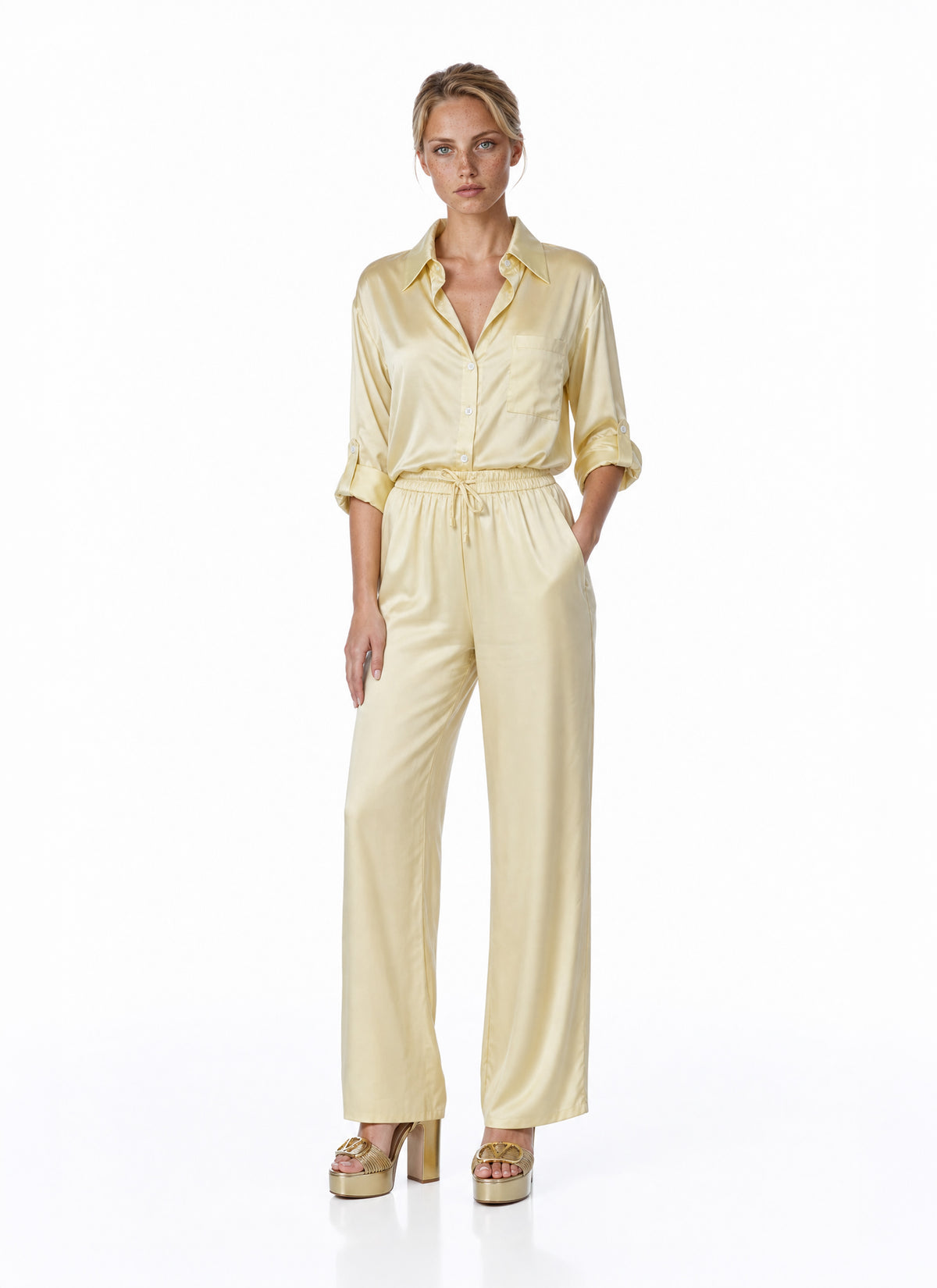 A woman modeling a pale yellow satin jumpsuit with a collar, rolled sleeves, and a drawstring waist, paired with metallic platform sandals, against a plain white studio background.