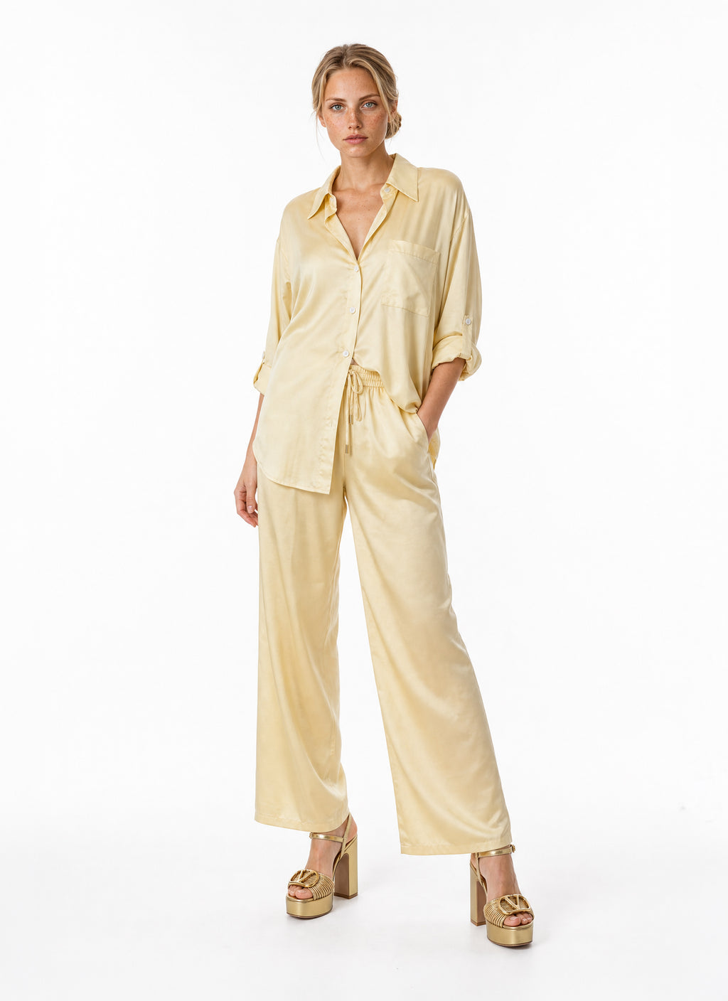 A woman modeling a pale yellow satin two-piece lounge set consisting of a long-sleeve button-up shirt and matching wide-leg pants, paired with gold platform heels against a plain white studio background.