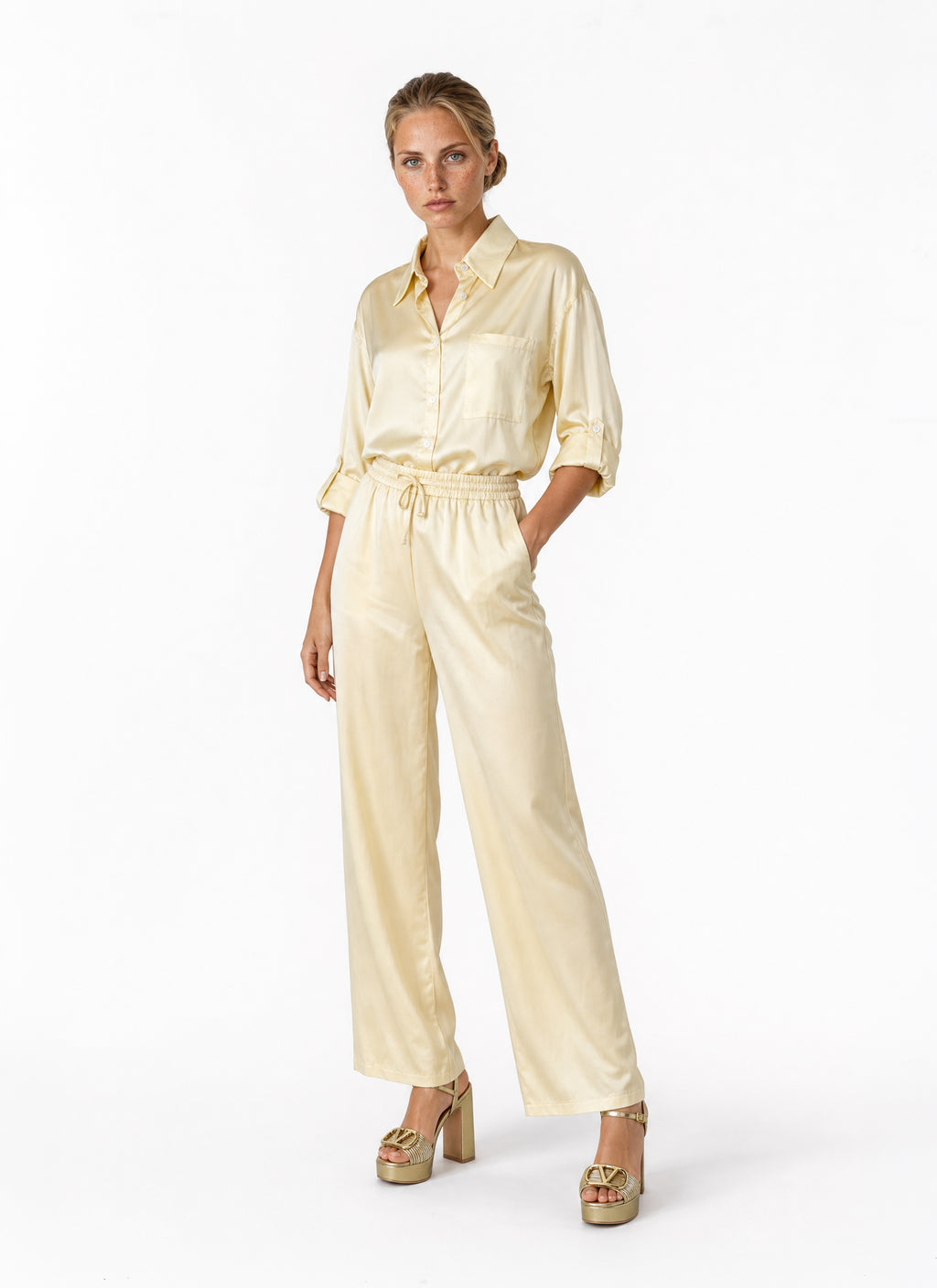 A woman modeling a pale cream satin blouse with a button-up shirt collar, rolled sleeves, a drawstring waist, and wide-leg trousers, paired with metallic gold platform heels against a plain white studio background.