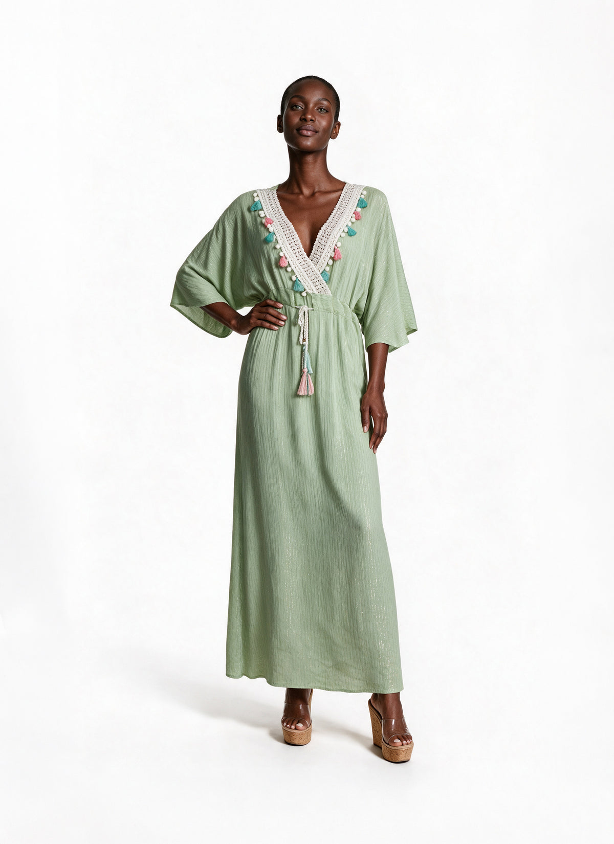 A woman modeling a sage green maxi kaftan dress with a white lace V-neckline, colorful embroidered details, and a matching drawstring waist, set against a plain white studio background.