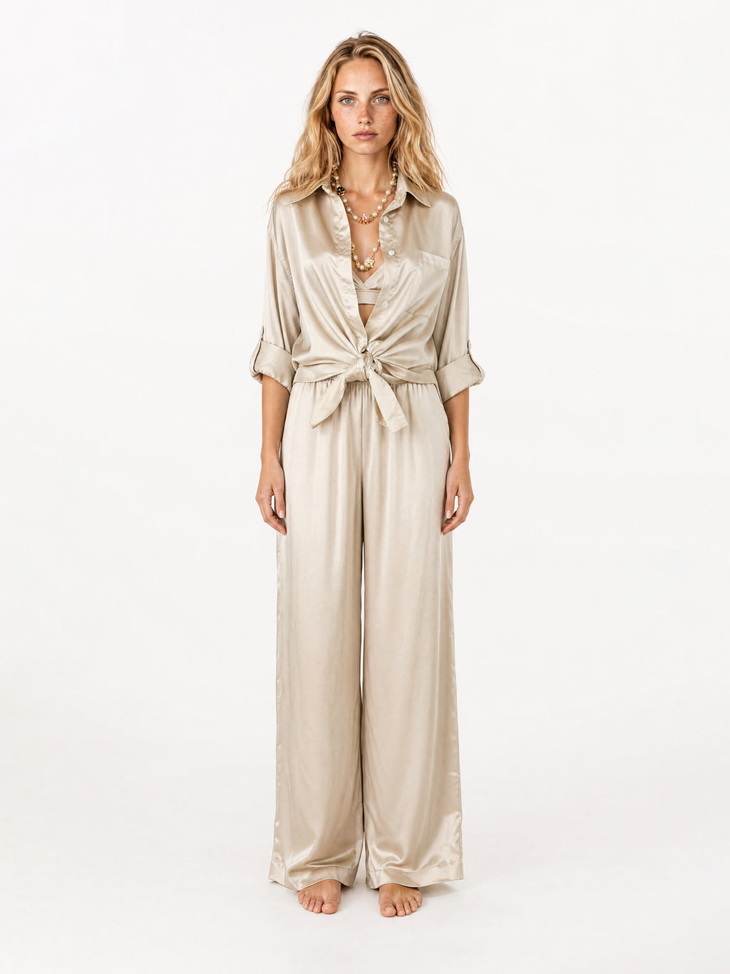 A woman stands against a plain white studio background wearing a shiny champagne-beige satin blouse with three-quarter sleeves, a tie-front waist, and wide legs.