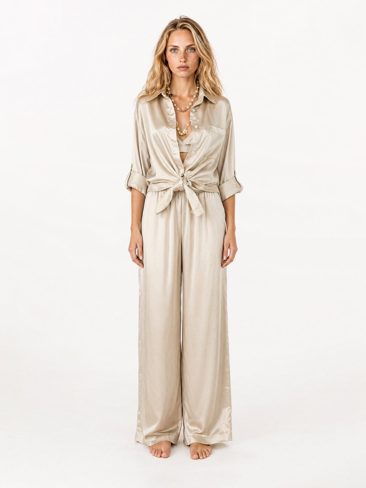 A woman stands against a plain white studio background wearing a shiny champagne-beige satin blouse with three-quarter sleeves, a tie-front waist, and wide legs.