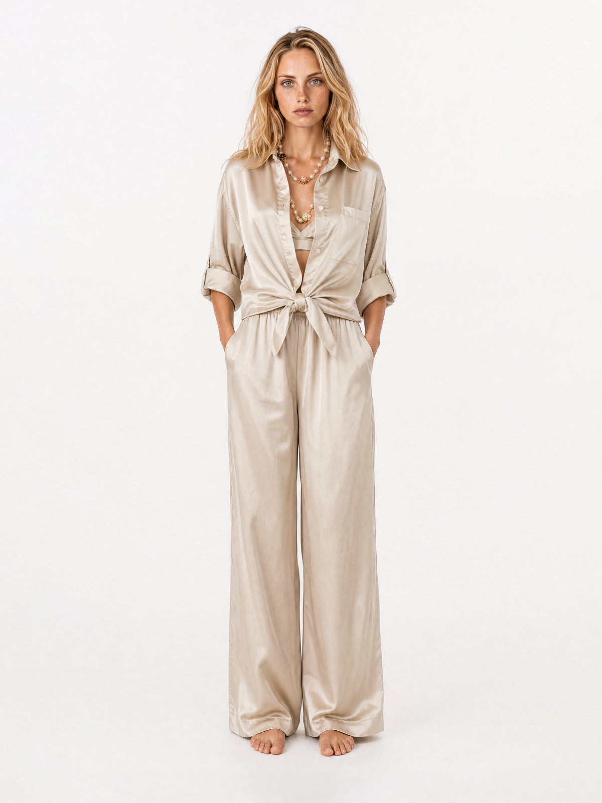 A woman stands barefoot against a plain white studio background wearing a light beige satin-blouse and a pant with a tie-front waist, long sleeves, and wide legs.