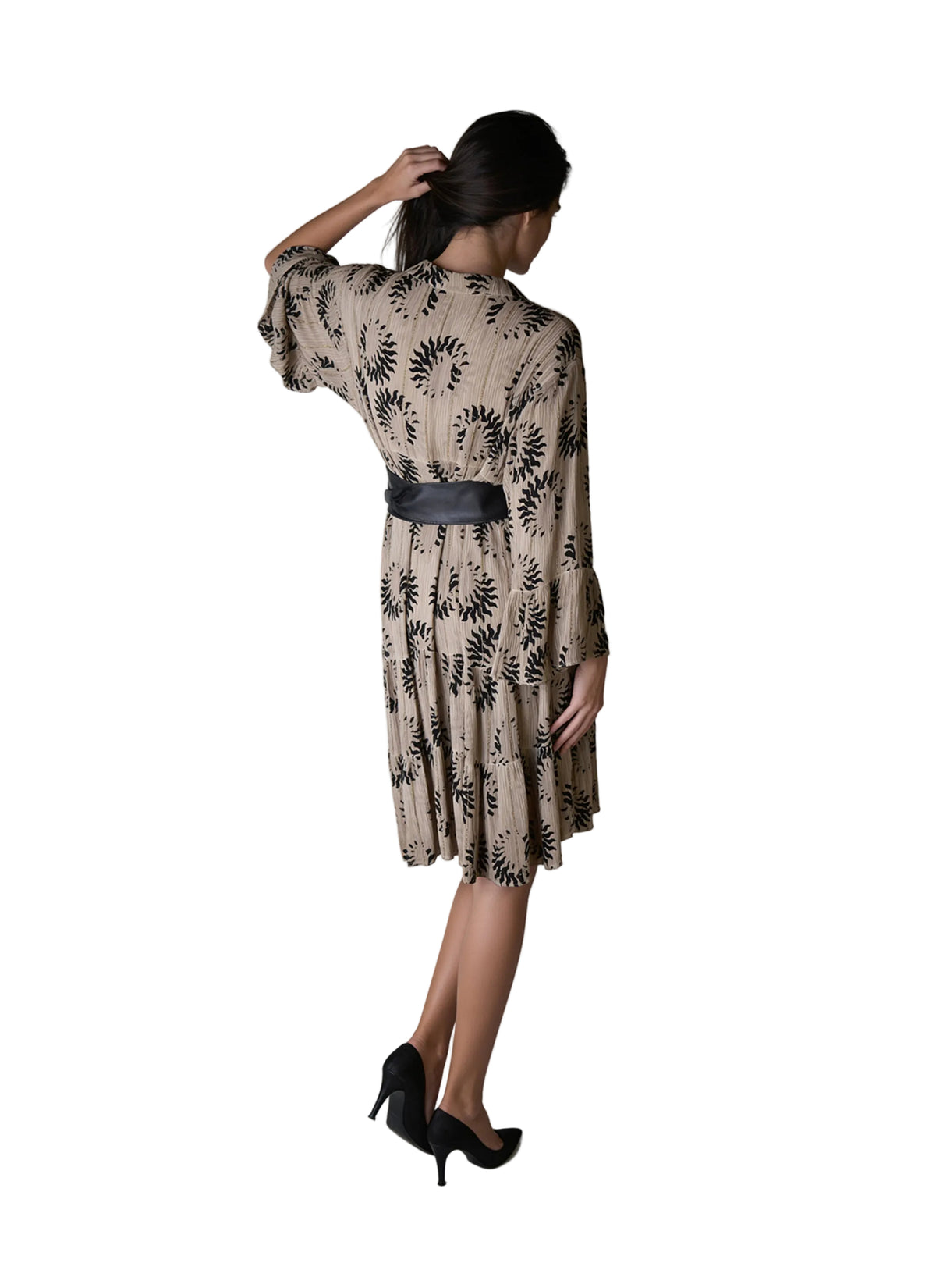 A woman is shown from behind wearing a beige midi-length dress with a black palm leaf pattern and a black belt, paired with black high heels, against a light gray studio background.