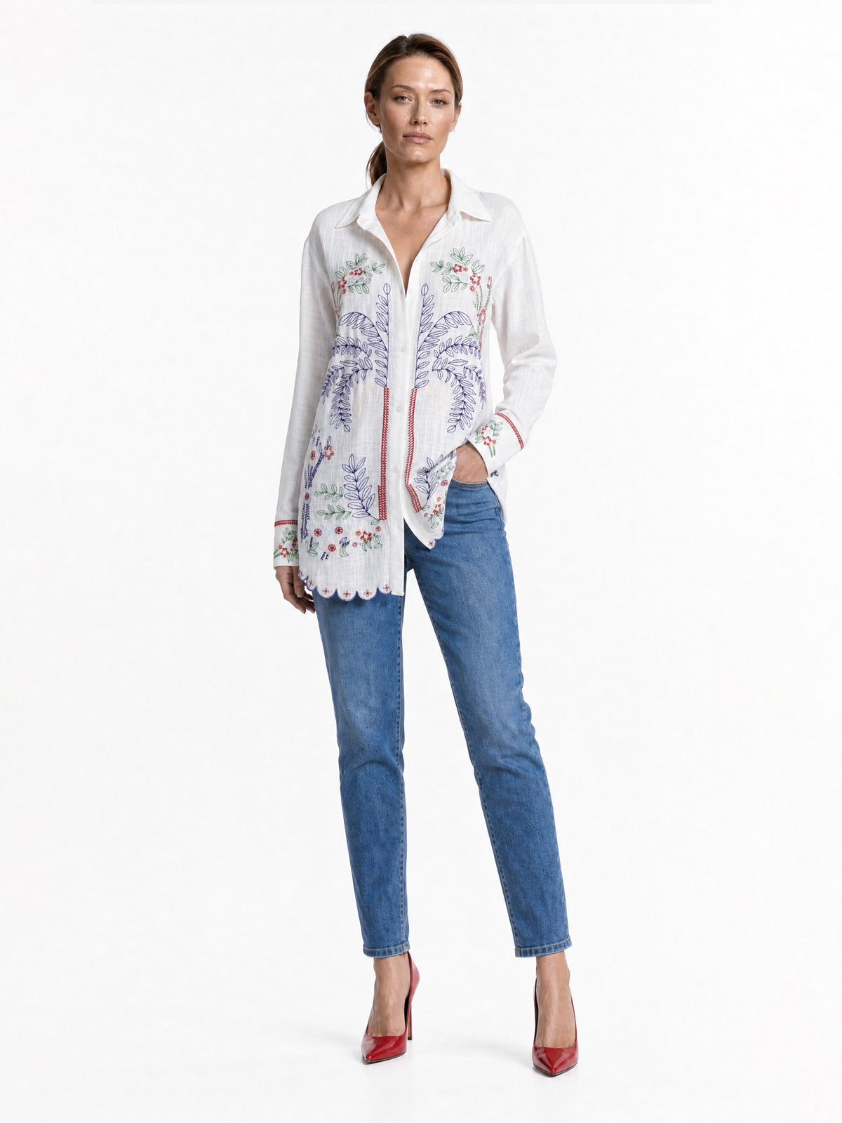A woman modeling a white long-sleeve button-up shirt with colorful floral embroidery, paired with blue jeans and red heels, against a plain white studio background.