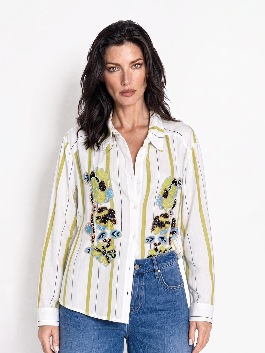 A woman modeling a white button-up shirt with vertical olive green stripes and colorful embroidered owl motifs on the front, paired with blue jeans against a plain white studio background.