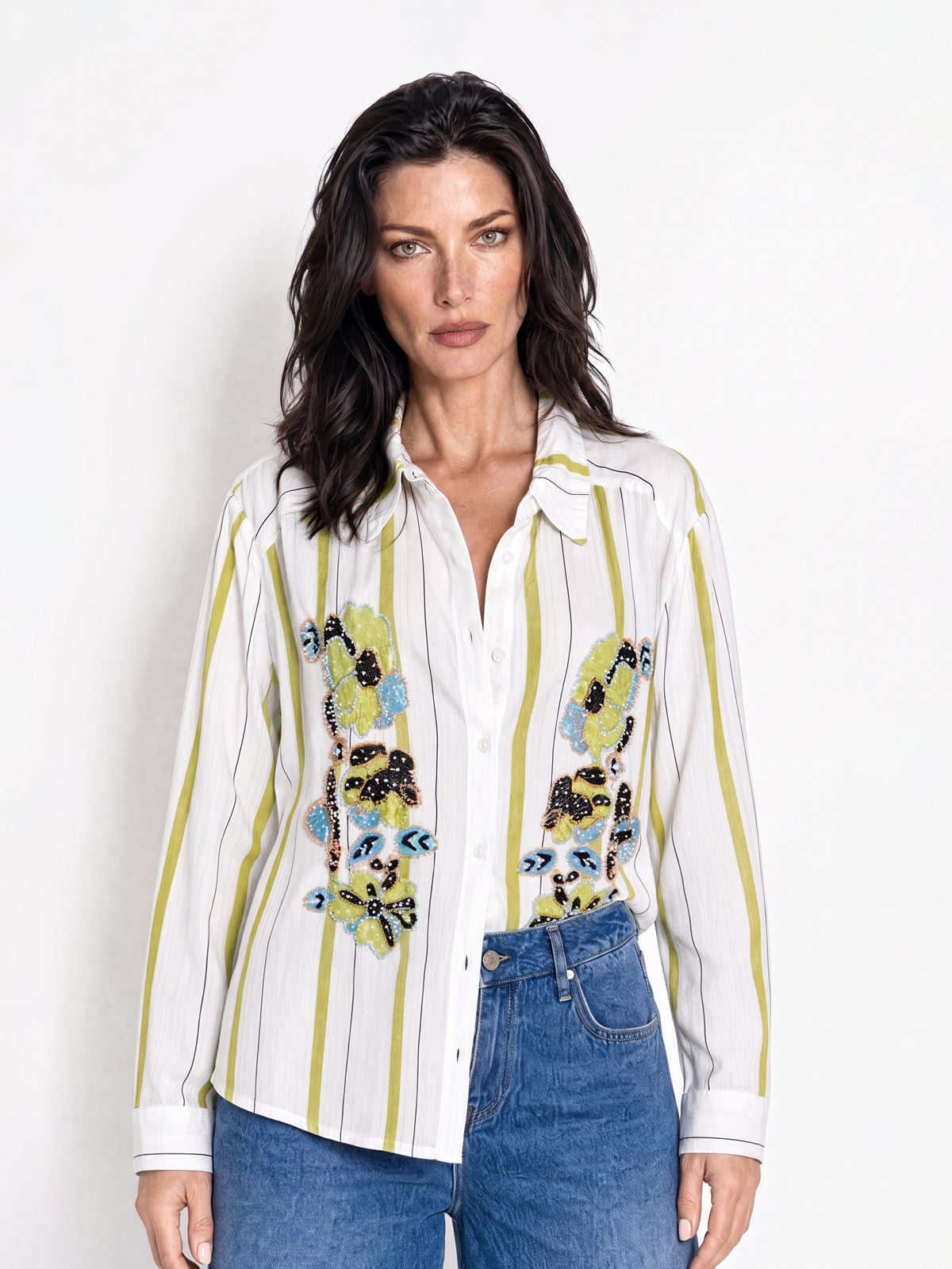 A woman modeling a white button-up shirt with vertical olive green stripes and colorful embroidered owl motifs on the front, paired with blue jeans against a plain white studio background.