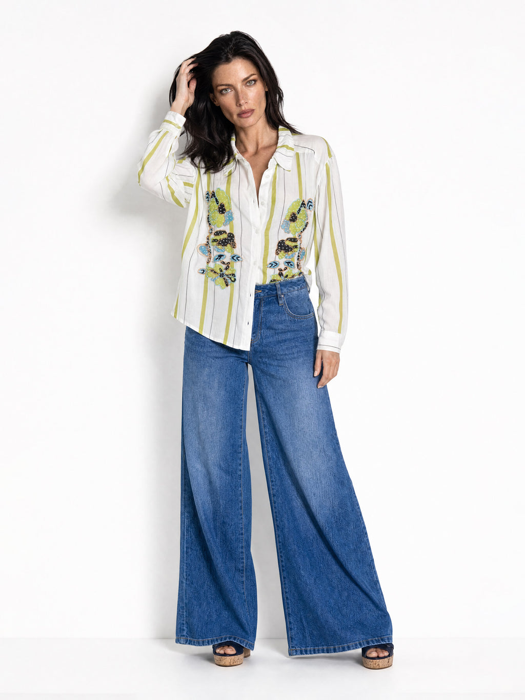 A model stands against a white studio background wearing a white long-sleeve button-up shirt with vertical yellow stripes and embroidered green and green floral details on the front, paired with high-waisted wide-leg blue jeans and brown sandals.