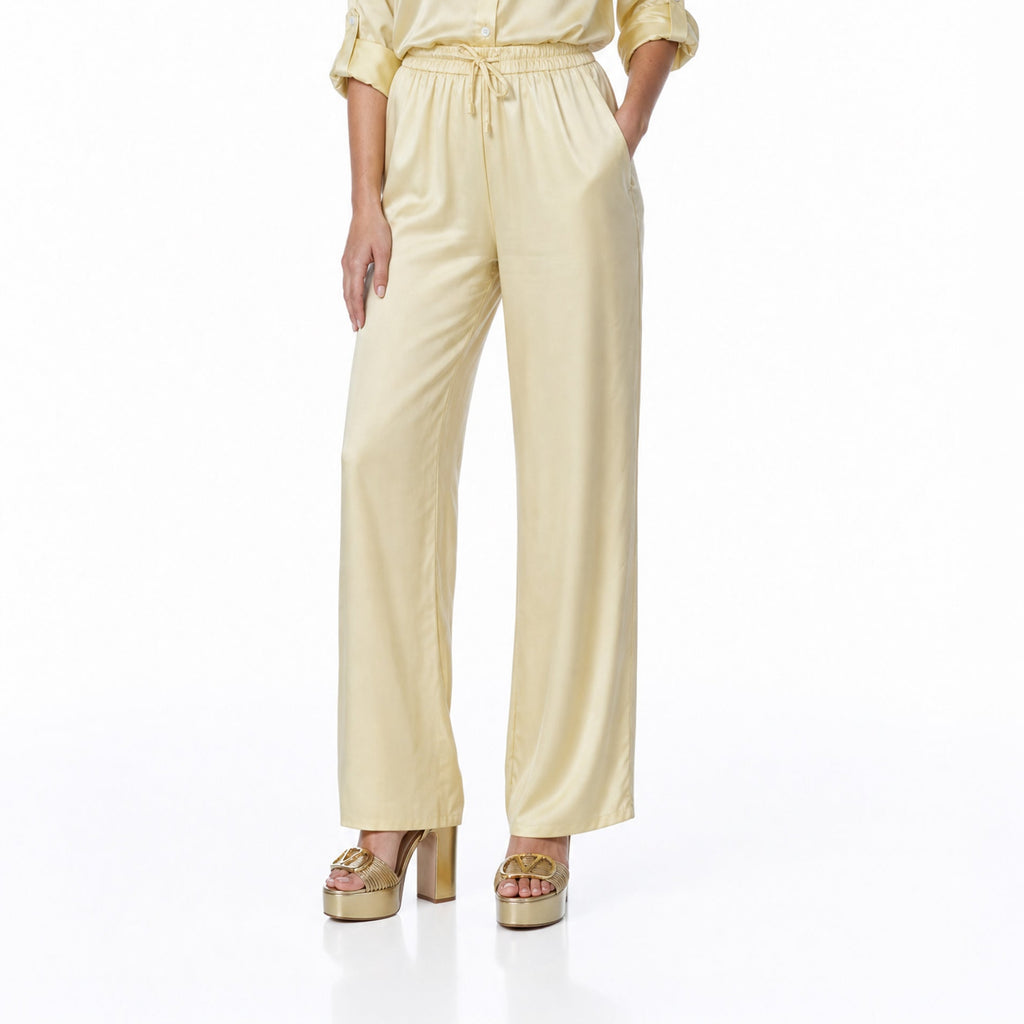 A woman modeling a pale yellow satin jumpsuit with a collar, rolled sleeves, and a drawstring waist, paired with metallic platform sandals, against a plain white studio background.