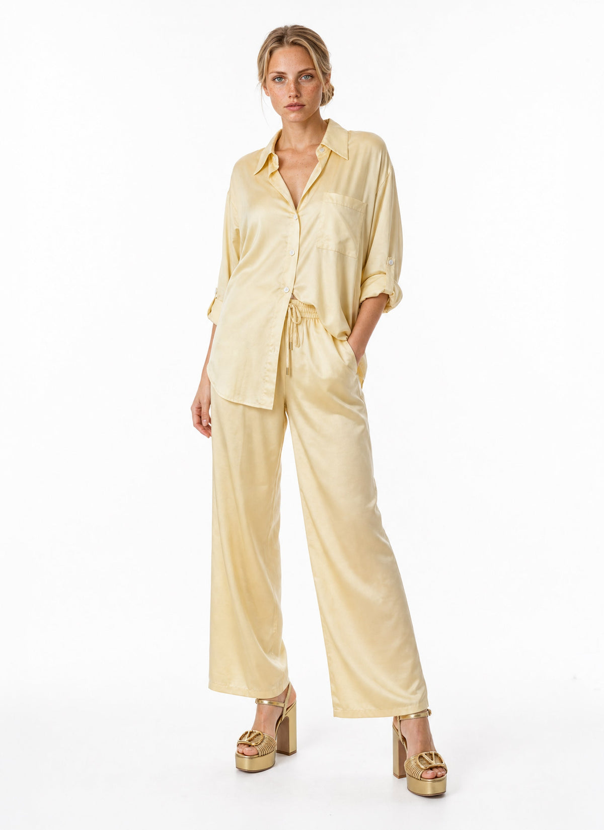 A woman modeling a pale yellow satin two-piece lounge set consisting of a long-sleeve button-up shirt and matching wide-leg pants, paired with gold platform heels against a plain white studio background.