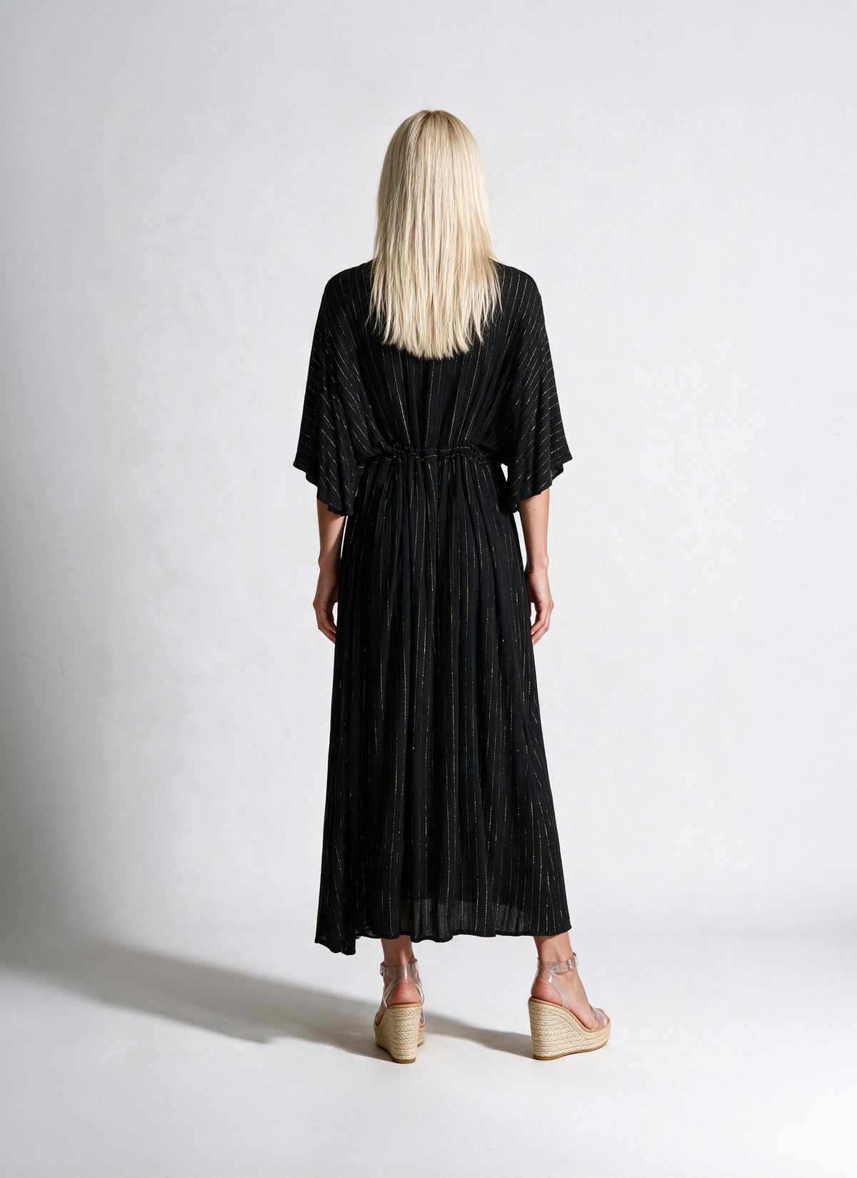 A woman viewed from behind wearing a black pleated kaftan dress with elbow-length sleeves and a tie waist, paired with beige wedge sandals, against a light gray studio background.