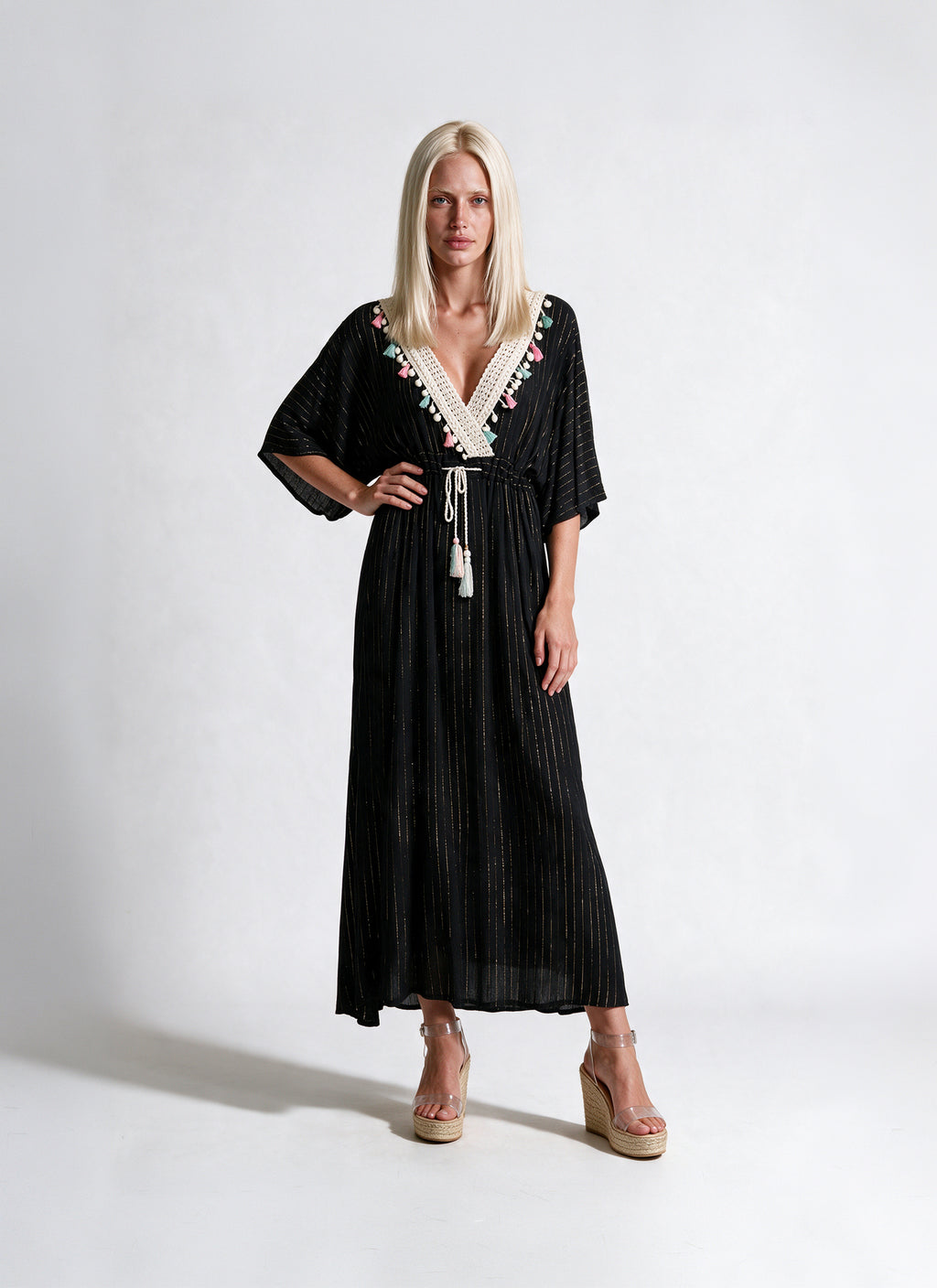 A blonde model wearing a black pleated maxi dress with a white beaded V-neckline and tassel tie, paired with beige wedge sandals, against a plain white studio background.