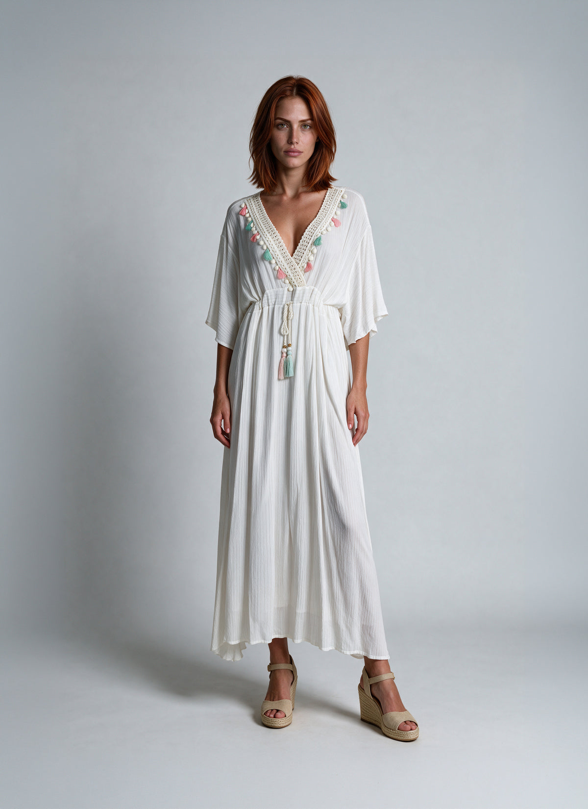 A woman stands against a light gray studio background wearing a flowing white maxi dress with a V-neckline, short wide sleeves, a tie waist, and colorful embroidered detailing along the neckline. She is also wearing beige wedge sandals.