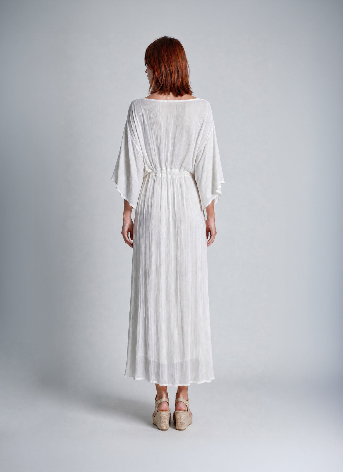 A woman with red hair is shown from the back wearing a white, loose-fitting midi dress with wide three-quarter sleeves and a gathered waist. She is standing against a neutral light gray studio background.