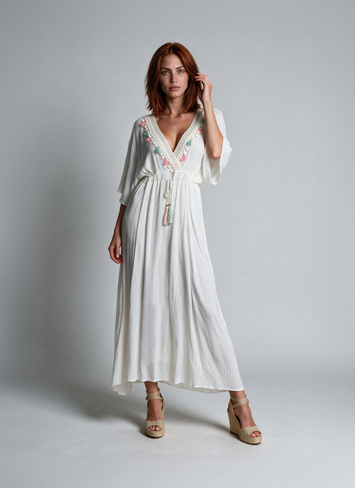 A woman modeling a white maxi dress with colorful floral embroidery along the V-neckline and waist tie, paired with beige wedge sandals against a light gray studio background.
