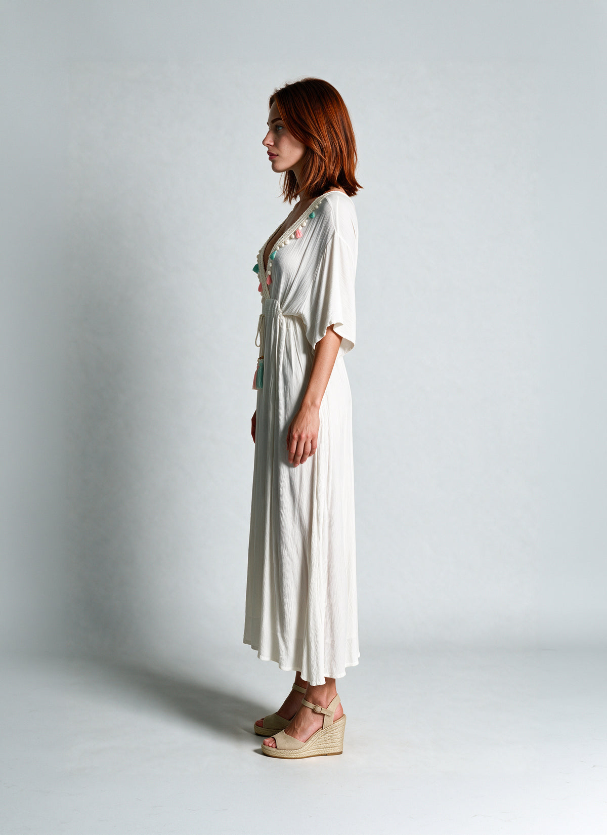 A woman stands in profile wearing a long white midi dress with short sleeves, a V-neckline, and delicate floral embroidery near the chest. She is wearing beige wedge sandals against a plain light gray studio background.