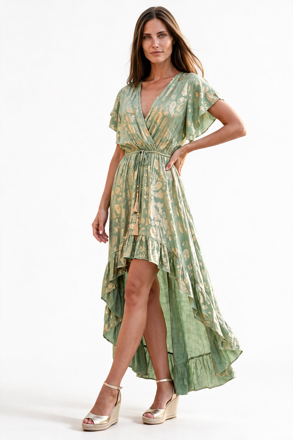 A woman modeling a light green and gold patterned high-low wrap dress with flutter sleeves and a tie waist, paired with beige wedge sandals, against a plain white studio background.