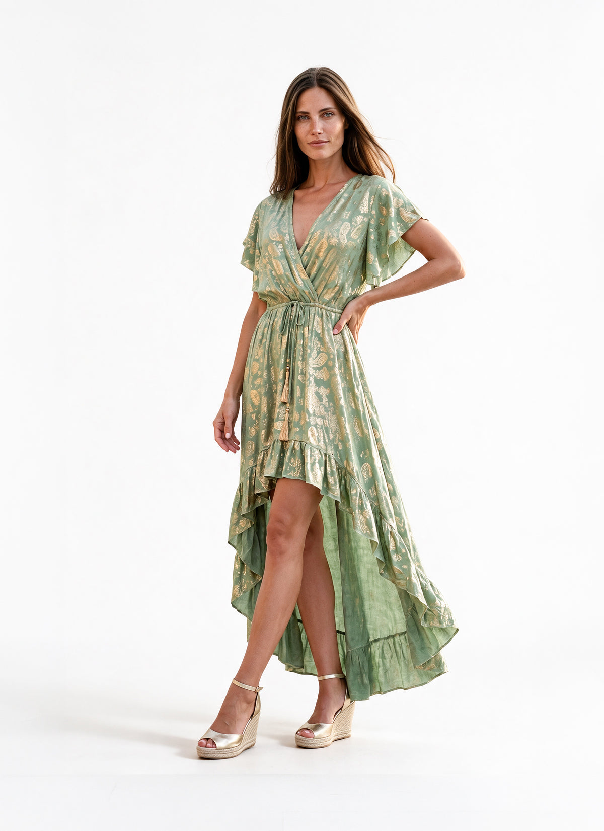 A woman modeling a light green and gold patterned high-low midi dress with flutter sleeves and a tie waist, paired with metallic gold wedge sandals, against a plain white studio background.
